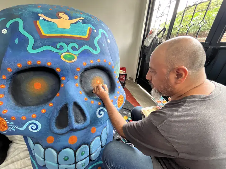 Artista pinta un cráneo azul con puntos y grecas en naranja.
